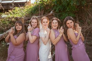 Bridal party poses together outdoors wearing matching mauve bridesmaid dresses with the bride in traditional white.