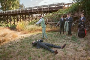 Playful wedding party staging a dramatic scene near rustic wooden bridge in outdoor setting.