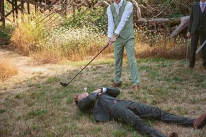 Two people in a rural field with someone in overalls holding a rake while another person lies on the ground.