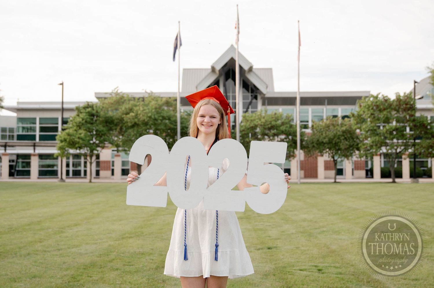 Graduate wearing white regalia and red cap holds large 2023 numbers while standing on green lawn in front of campus building.