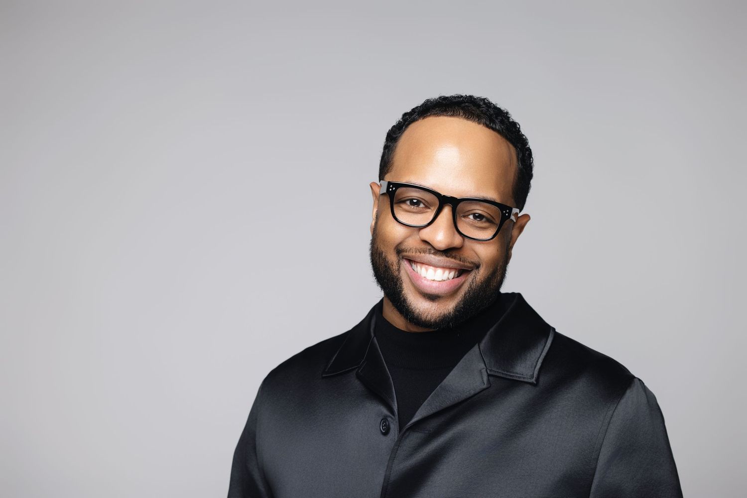 Professional headshot against a gray background showing person in black turtleneck sweater and glasses.