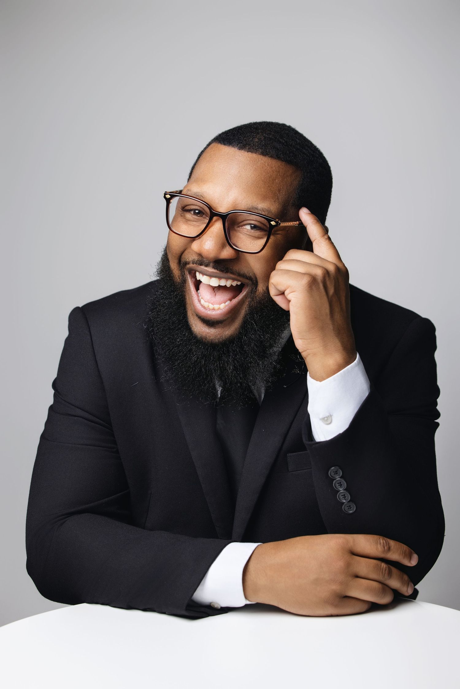 Professional in a black suit and glasses smiles joyfully while leaning on white surface in studio portrait.