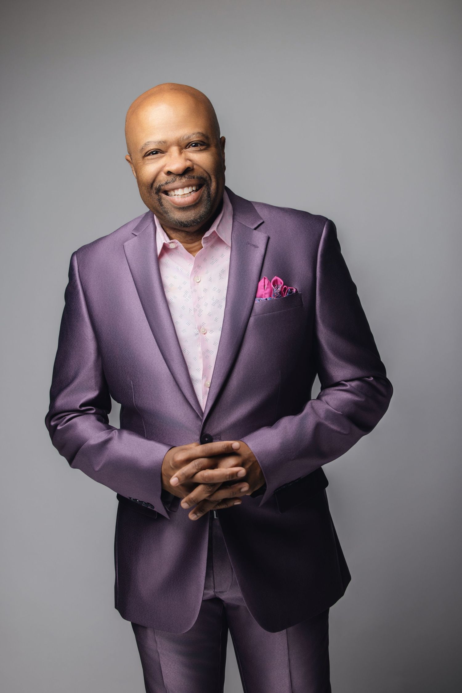 Person wearing purple suit with pink shirt and pocket square poses against gray backdrop.