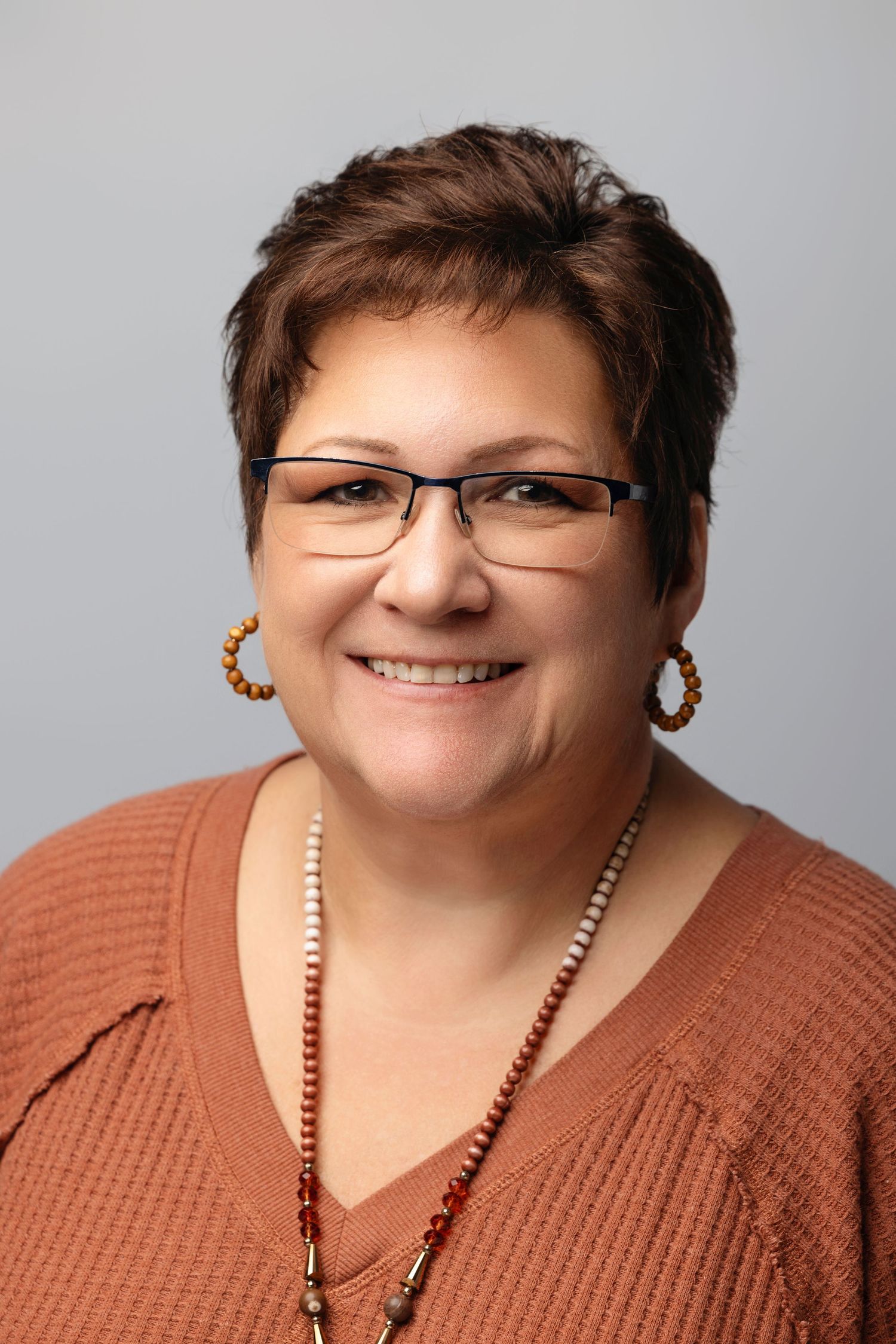 Close-up portrait of someone in rust-colored sweater and pearl necklace against gray background.