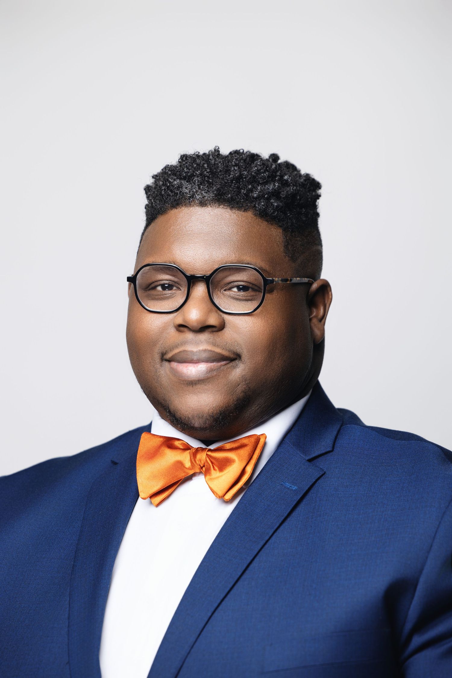 Person in navy suit with orange bow tie and glasses smiles warmly in professional headshot.