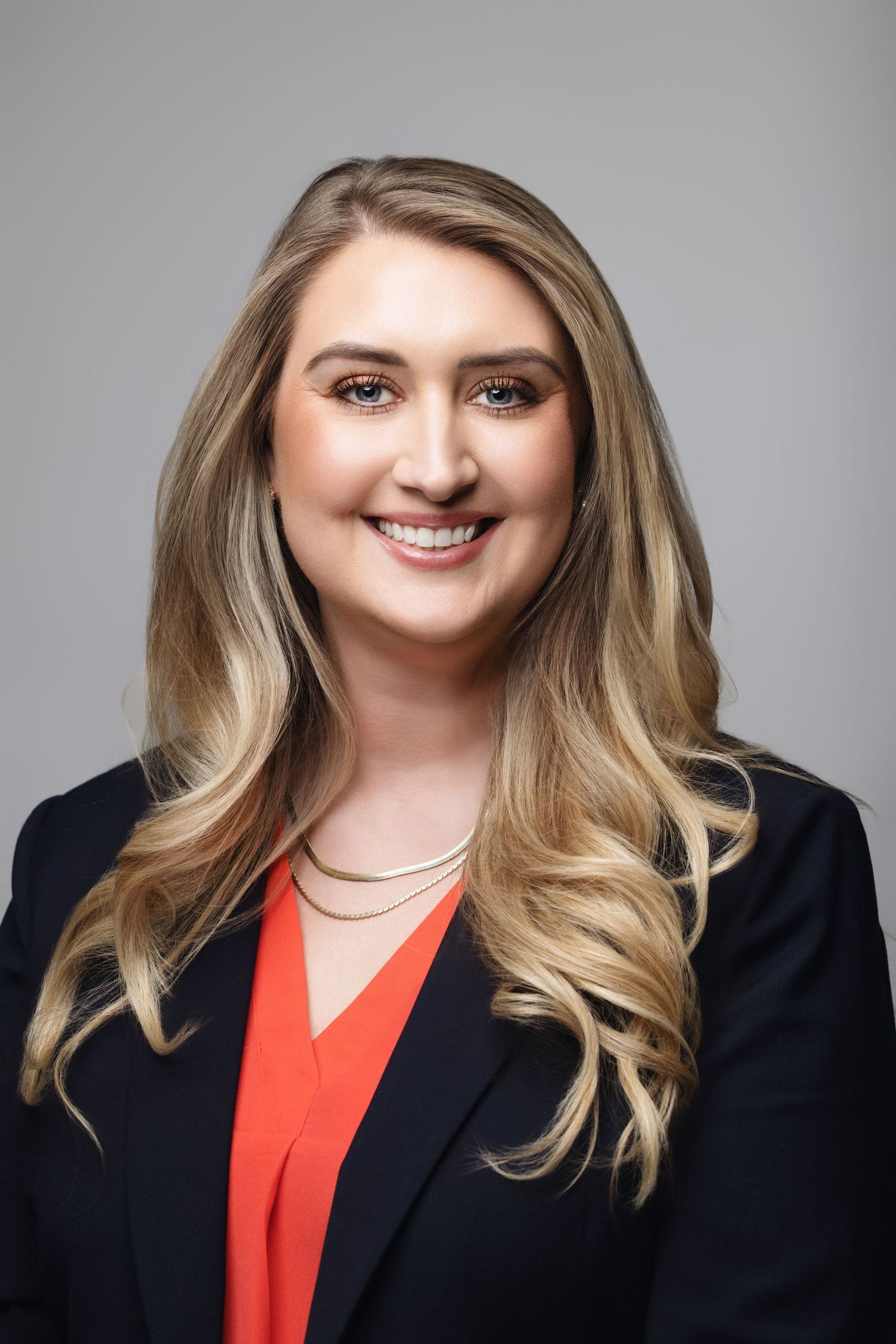 Professional portrait of someone in black blazer and coral top with long blonde hair.