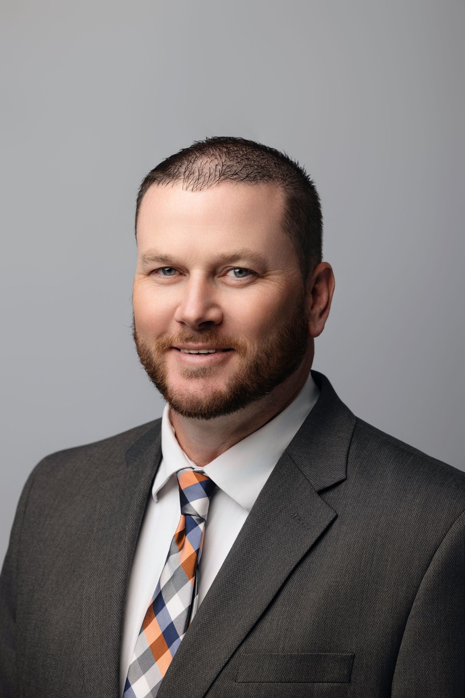Professional headshot of person in dark suit with striped tie against gray backdrop.