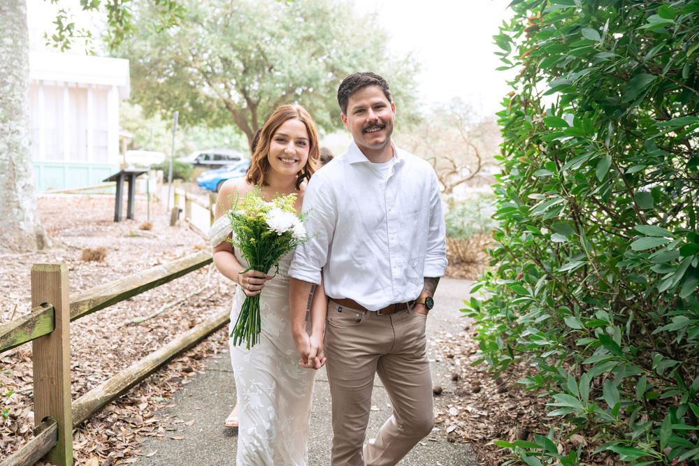 Couple in light-colored attire holding hands and walking through a lush garden path on a sunny day.