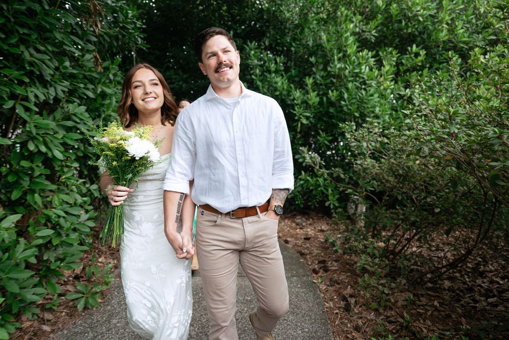 A couple in wedding attire walks through a lush forest path in summer.