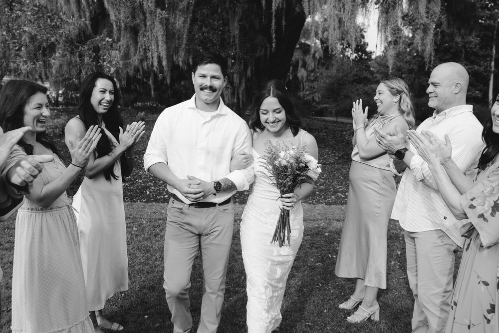 A joyful wedding party laughs together outdoors in black and white, with bridesmaids in light dresses beside a couple.