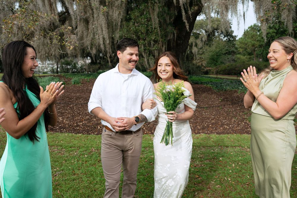 A group wearing white and green outfits celebrates together in a lush outdoor garden setting with Spanish moss trees.