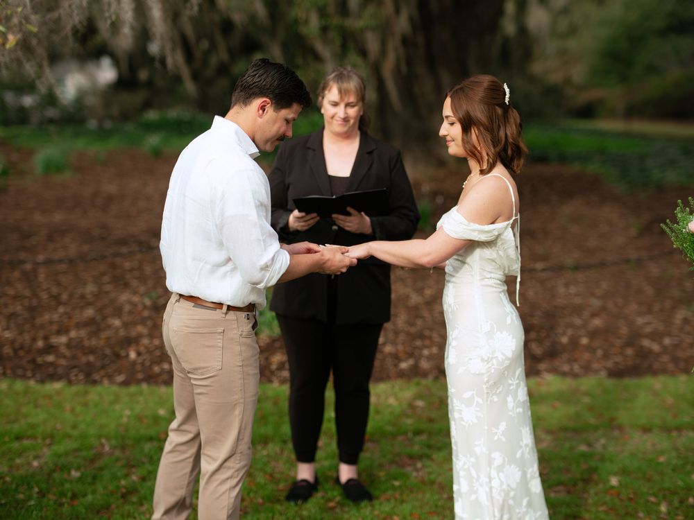 An intimate outdoor wedding ceremony takes place in a wooded area with an officiant and couple exchanging rings.
