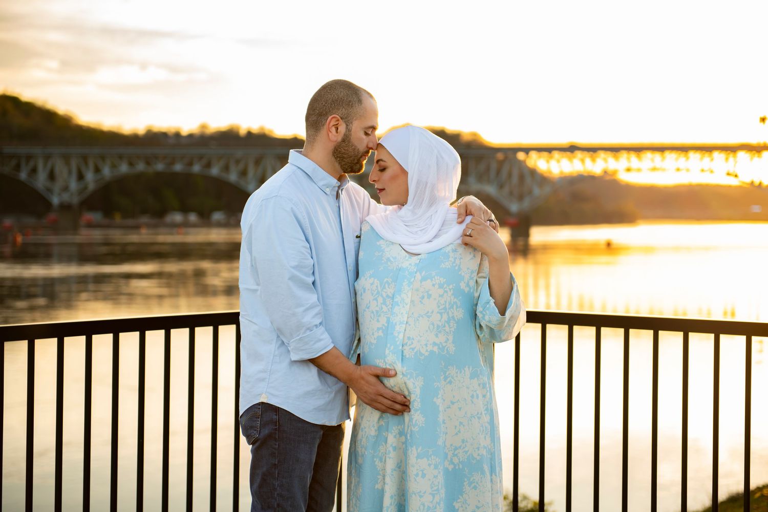 Couple embracing by a waterfront railing during golden sunset hour with cityscape in background.