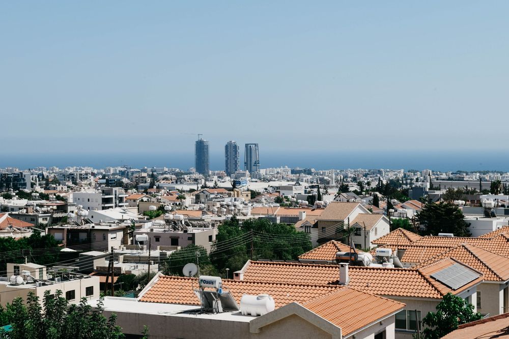 A panoramic cityscape view of a modern city with tall buildings and orange tiled rooftops against a blue sky and coastline.