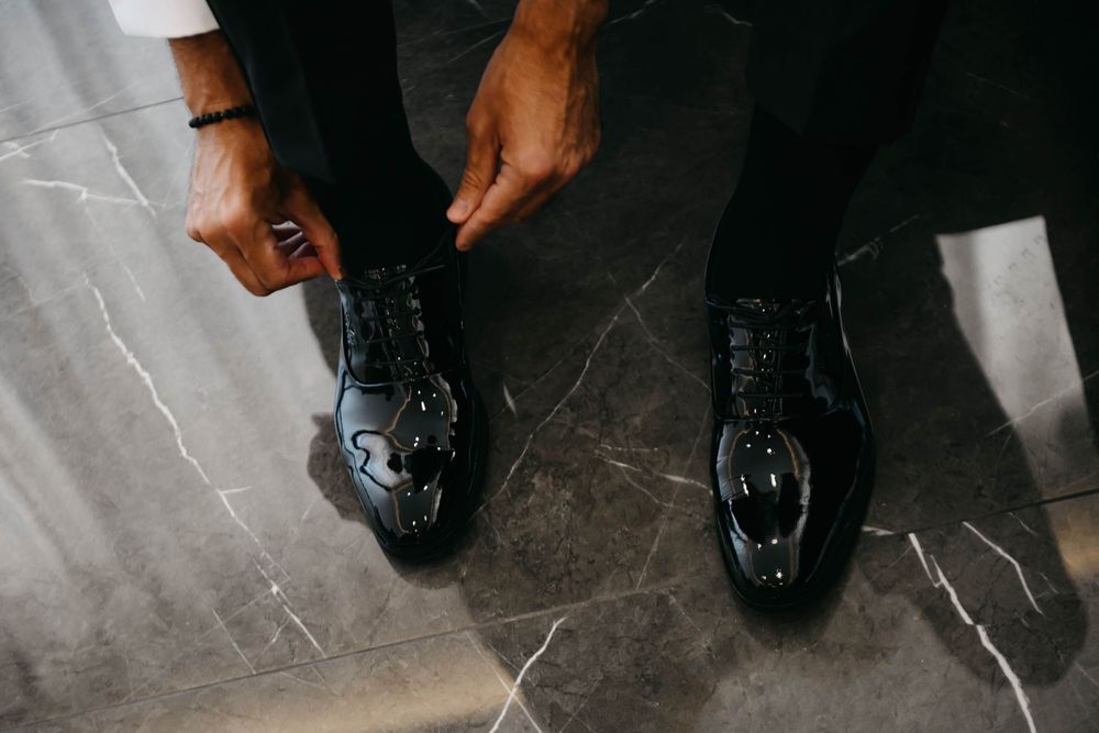 Hands tying laces on shiny black formal dress shoes against a polished marble floor.