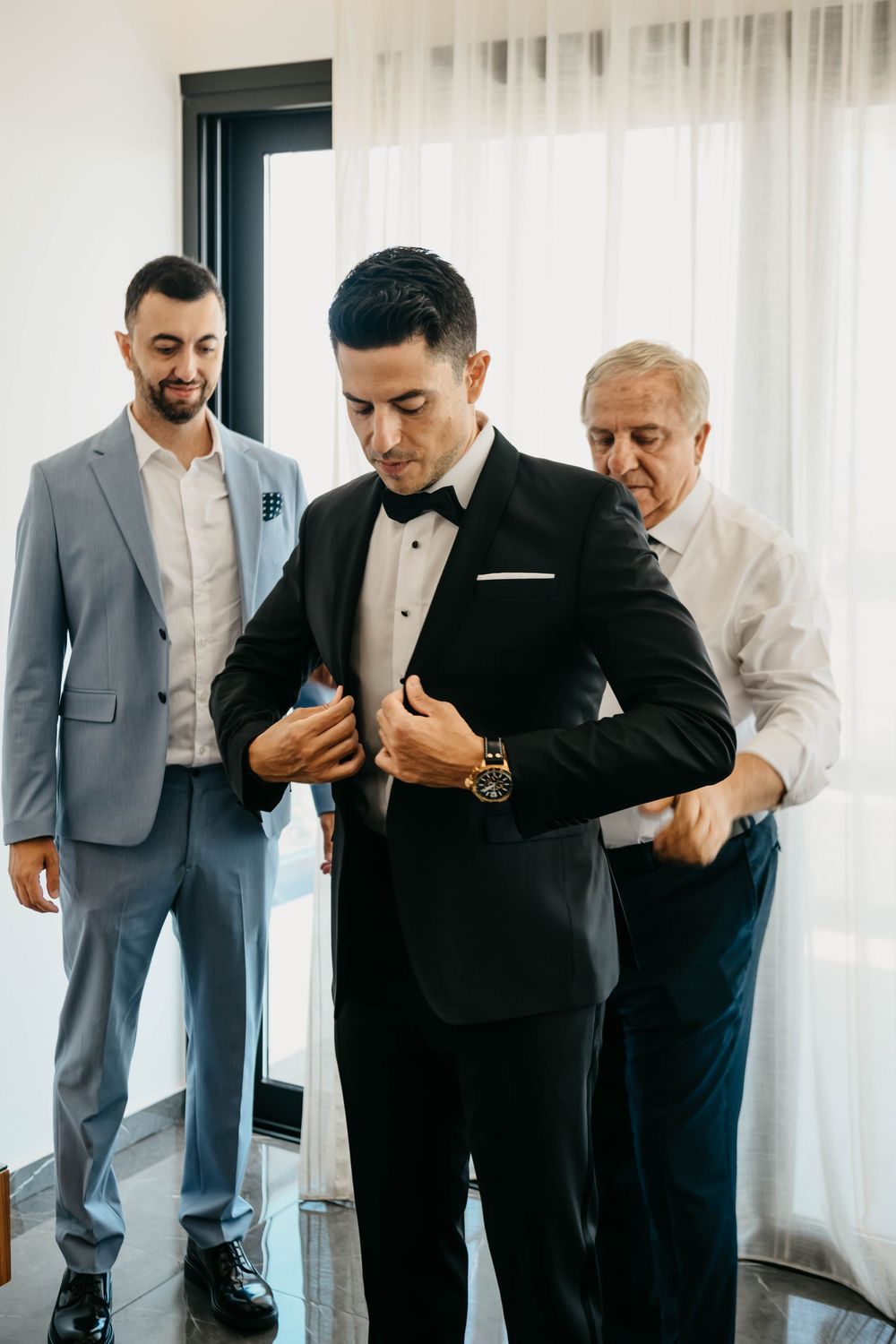 Men in formal attire prepare for a special occasion as groomsmen assist with final adjustments in a bright room.