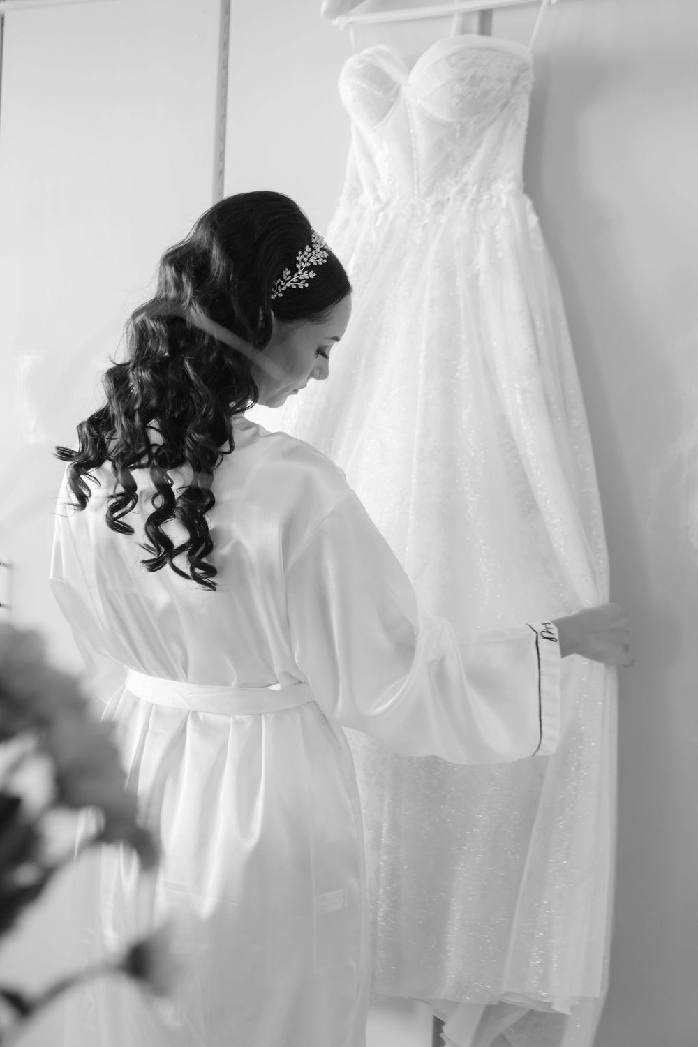 A bride in a white robe looking at her hanging white strapless wedding gown in a contemplative black and white photo.
