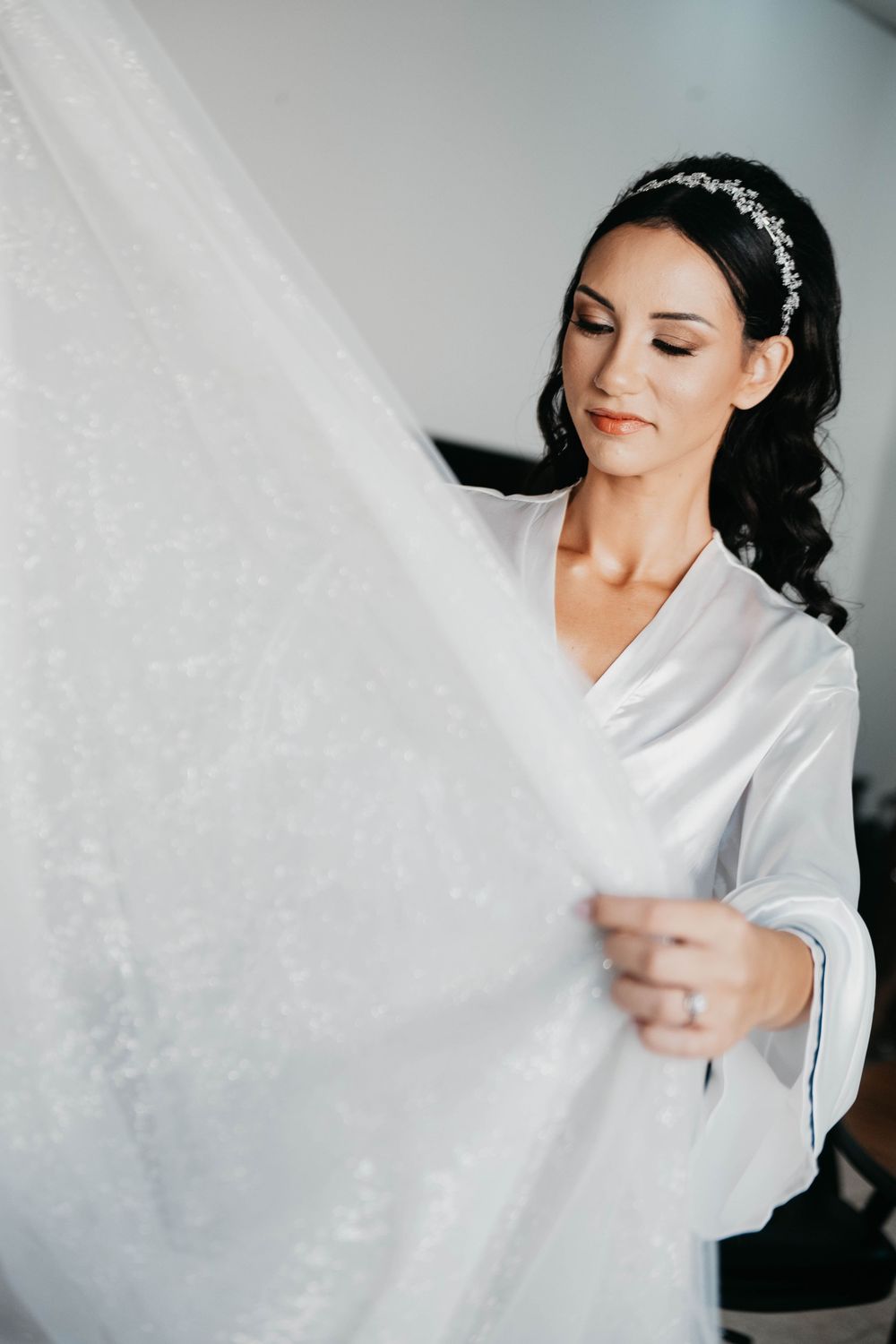 A bride in a white silk robe and crystal headpiece gently touches her wedding dress during wedding preparations.