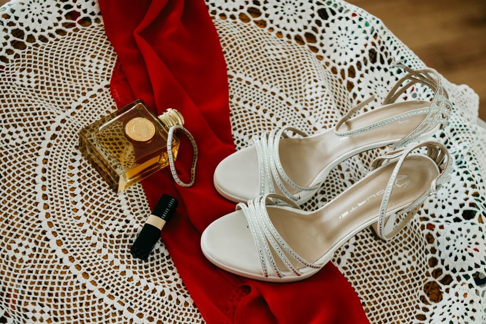 Elegant white bridal shoes and wedding rings displayed on vintage lace doily with red fabric accent.