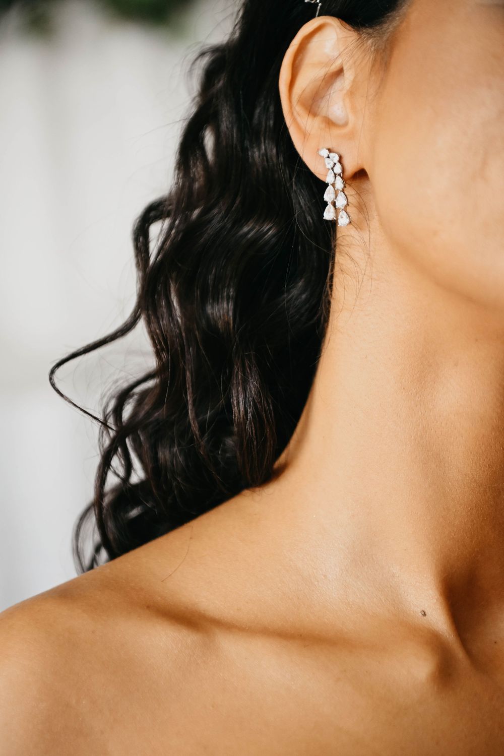 Close up of elegant diamond drop earring on ear with dark wavy hair and bare shoulder visible.