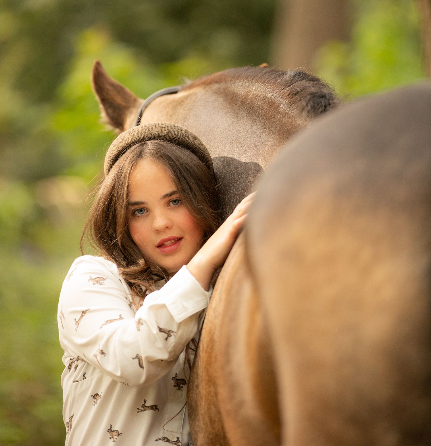 Someone wearing a white patterned top lovingly strokes a brown horse in a sunny outdoor setting.