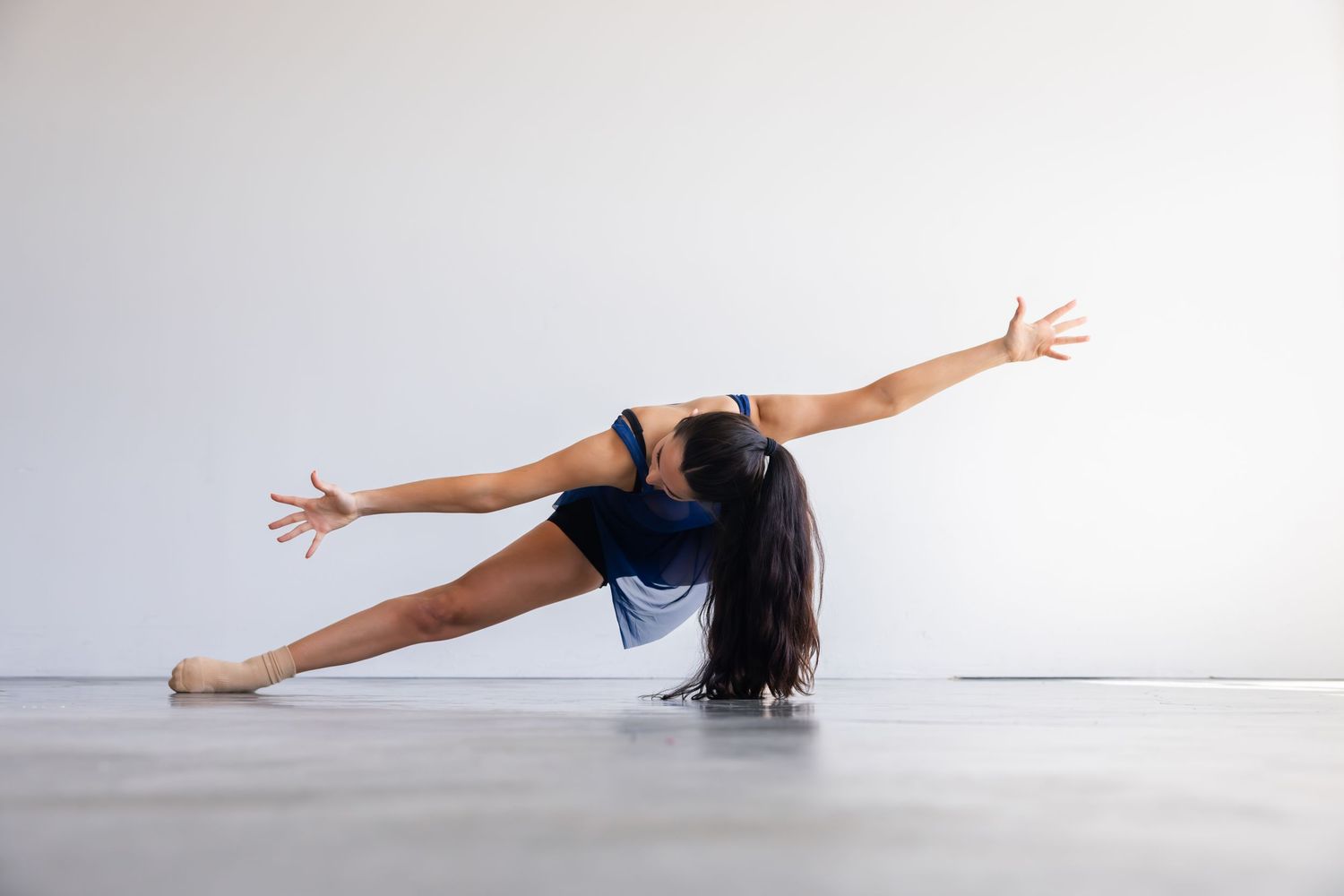 Dancer in blue leotard performs graceful contemporary dance move on studio floor against white wall.