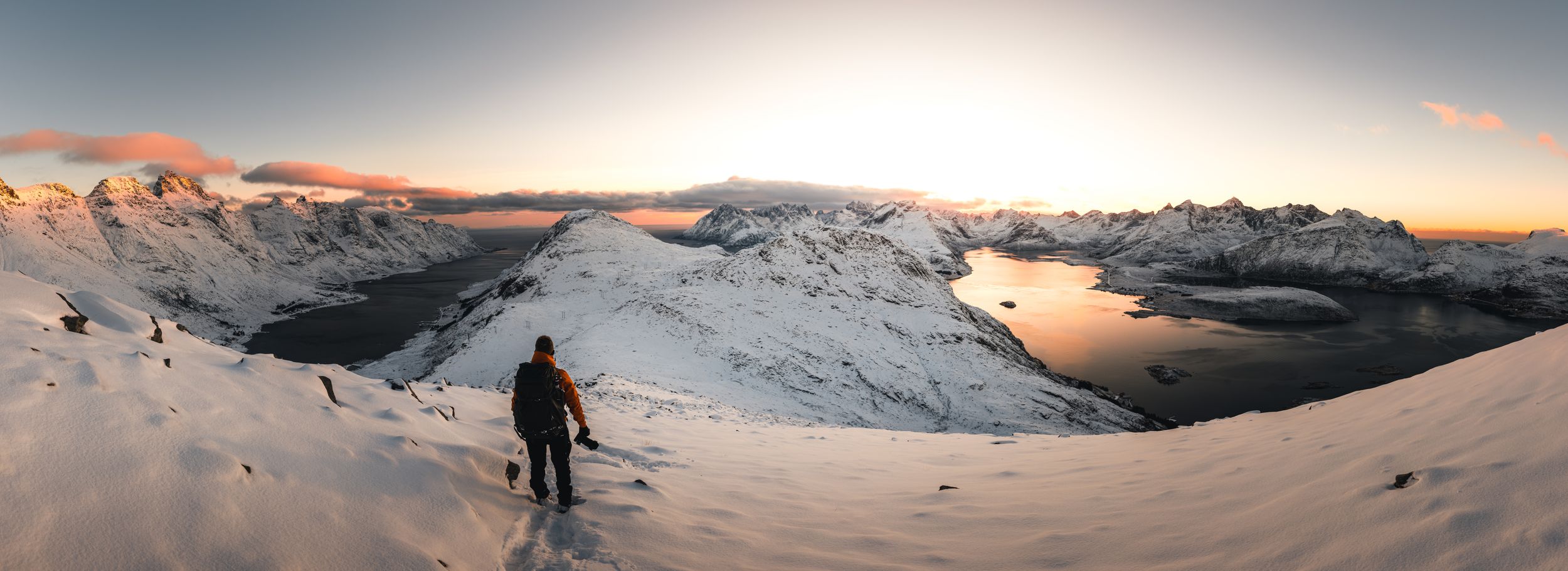A panoramic winter landscape view of snow-covered mountains illuminated by warm sunset light.