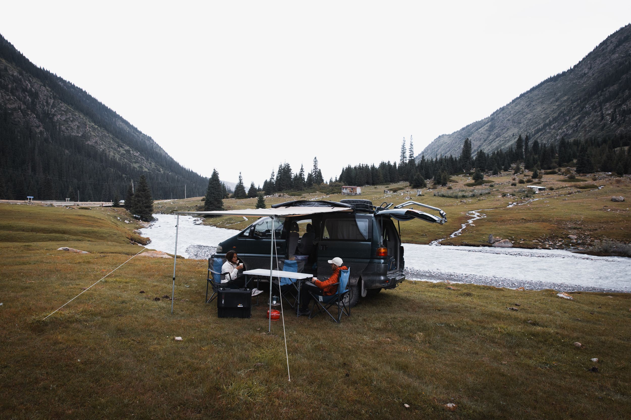 Black SUV parked at a scenic mountain campsite with melting snow patches and winding river in a remote valley setting.