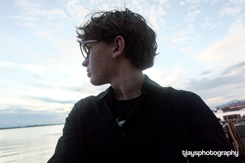 Young man with glasses gazes at waterfront scenery during golden hour outdoors.