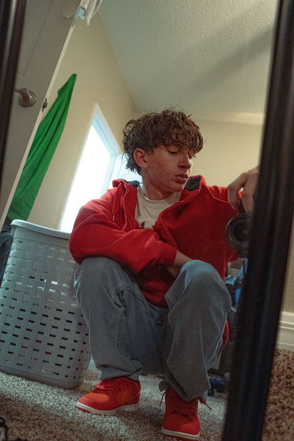 Young man in red hoodie sits on floor photographed through a mirror reflection.