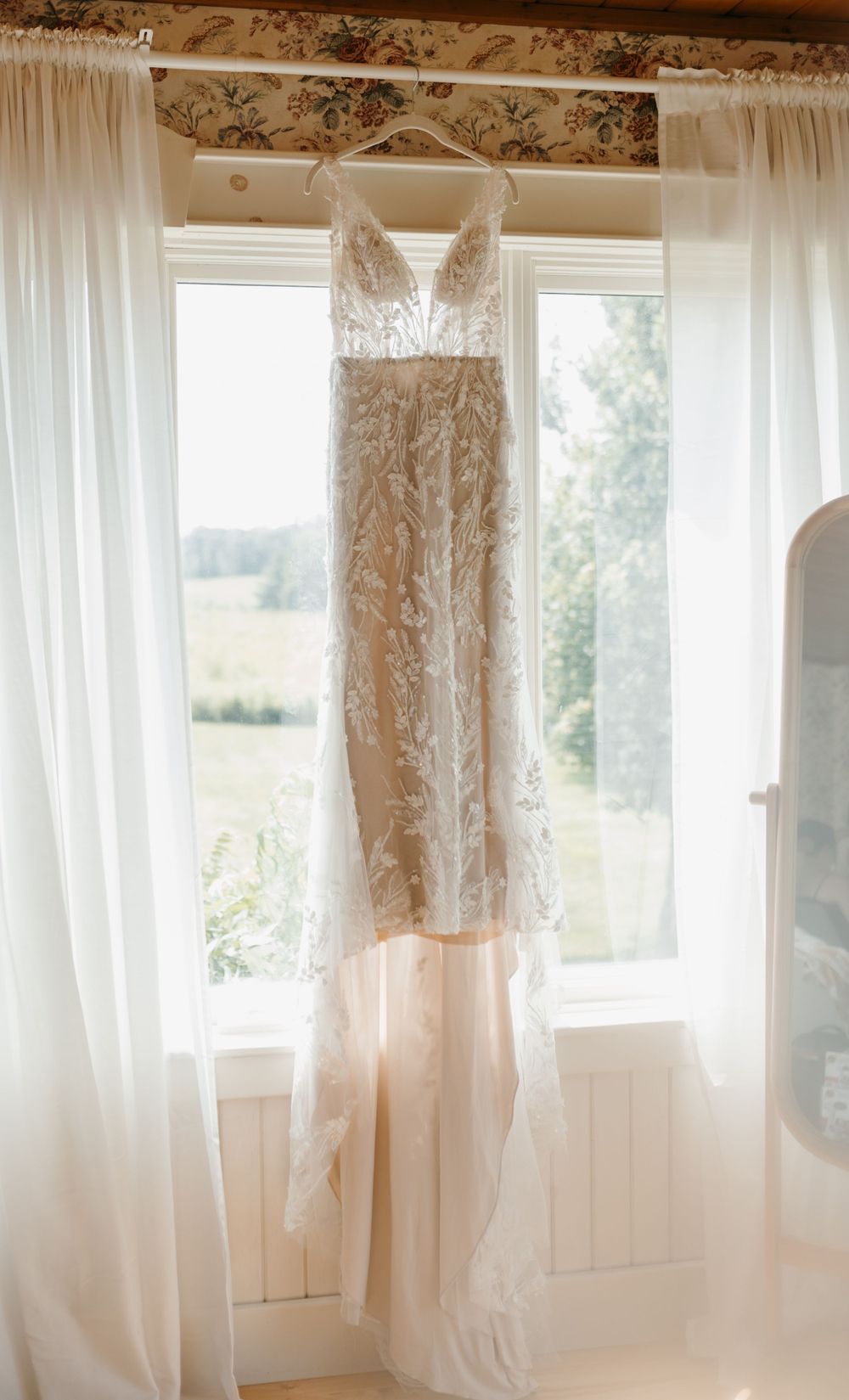 Lace wedding gown hanging in window with scenic green countryside view.