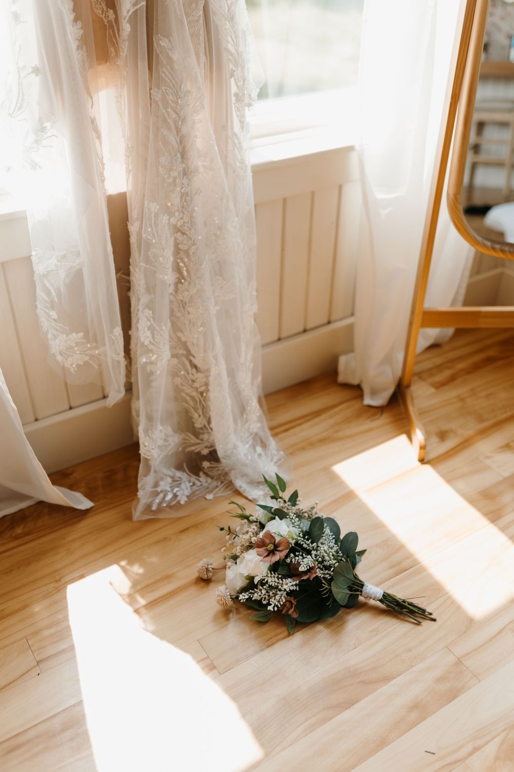Bridal bouquet with white roses and eucalyptus on hardwood floor beside lace wedding gown.