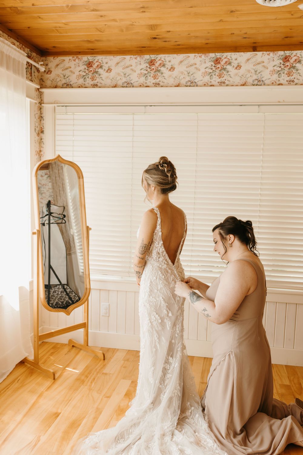Bridesmaid kneeling to help bride into lace wedding gown beside a gold floor mirror.