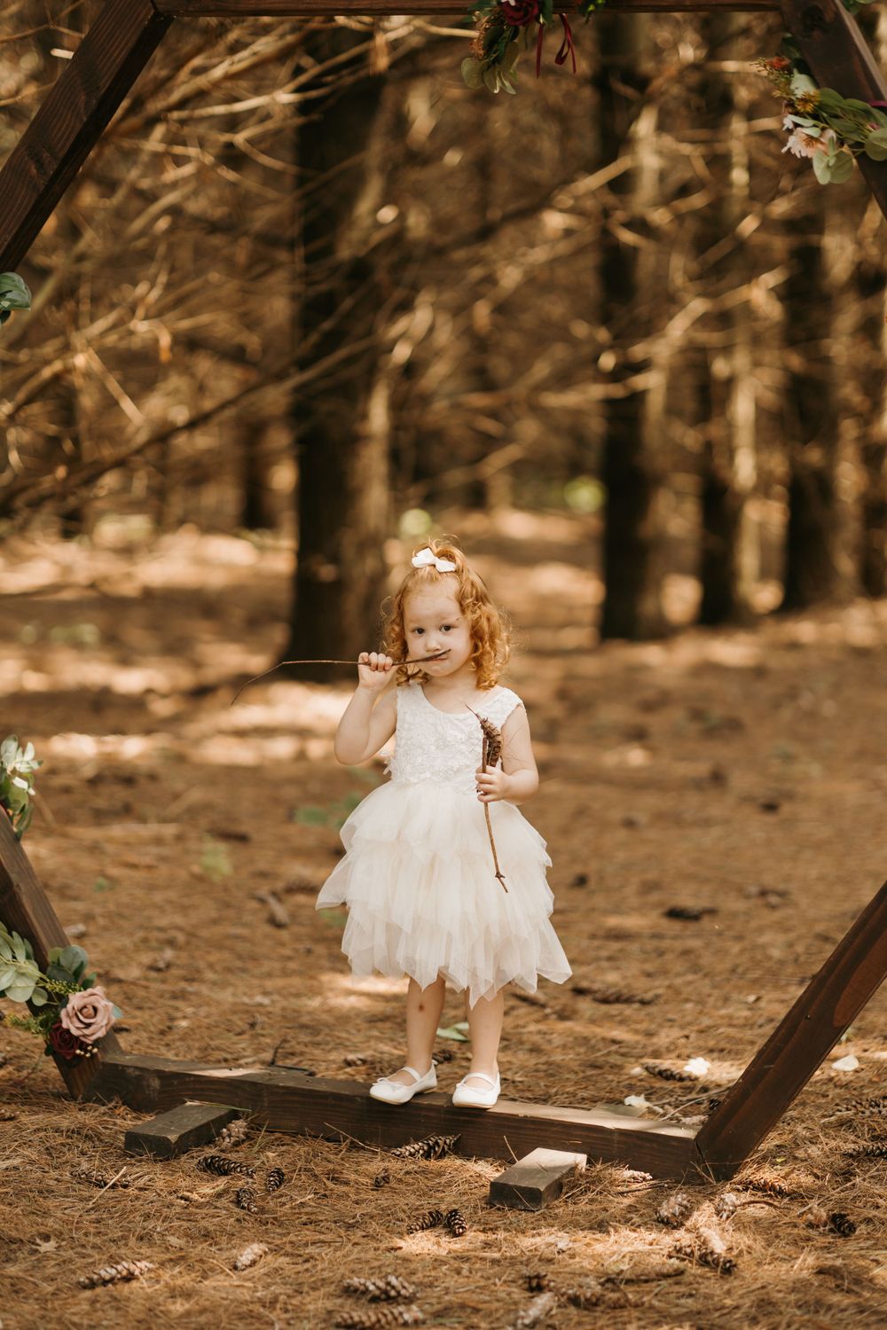 Adorable redhead flower girl in white tulle dress standing inside wooden hexagon wedding arch.