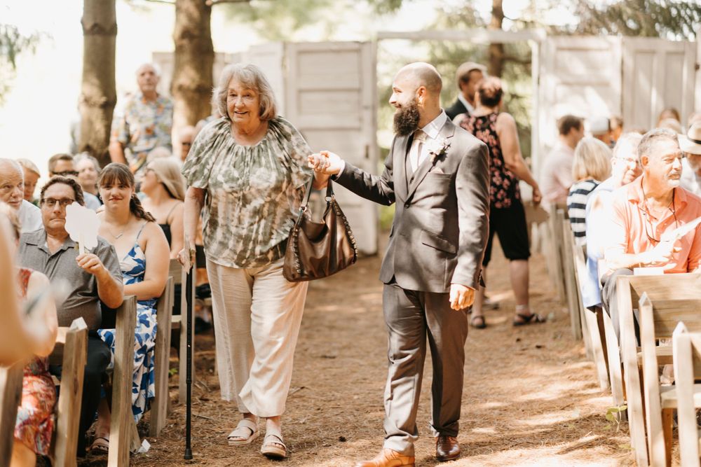 Groom in gray suit escorts elderly woman down outdoor woodland wedding ceremony aisle.