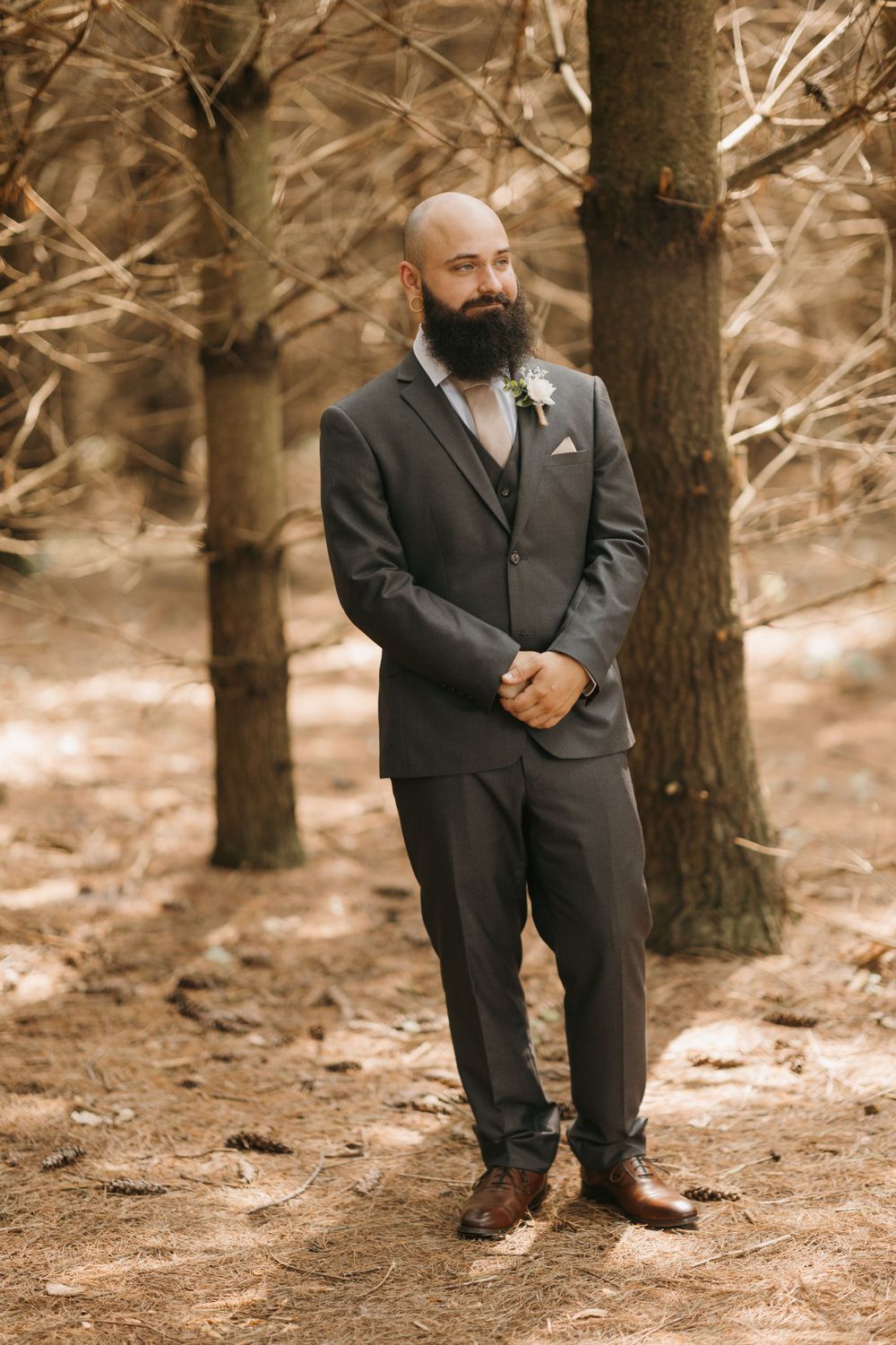 Handsome bald groom with beard in gray three-piece suit standing among trees at outdoor wedding.