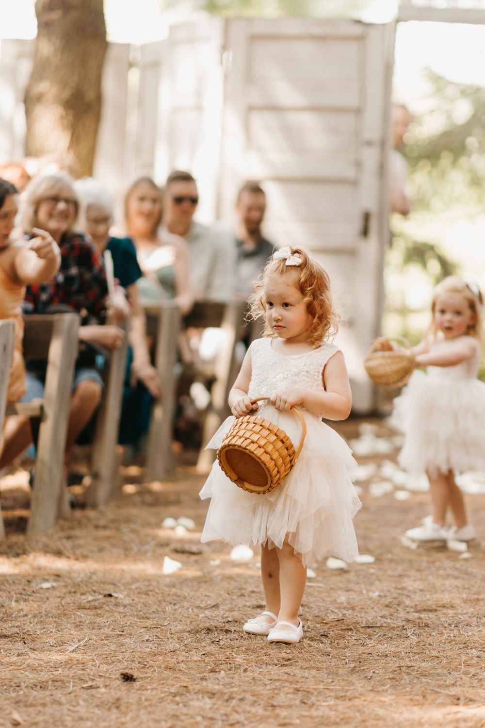 Curly-haired flower girl in white dress carrying wicker basket down outdoor wedding aisle.