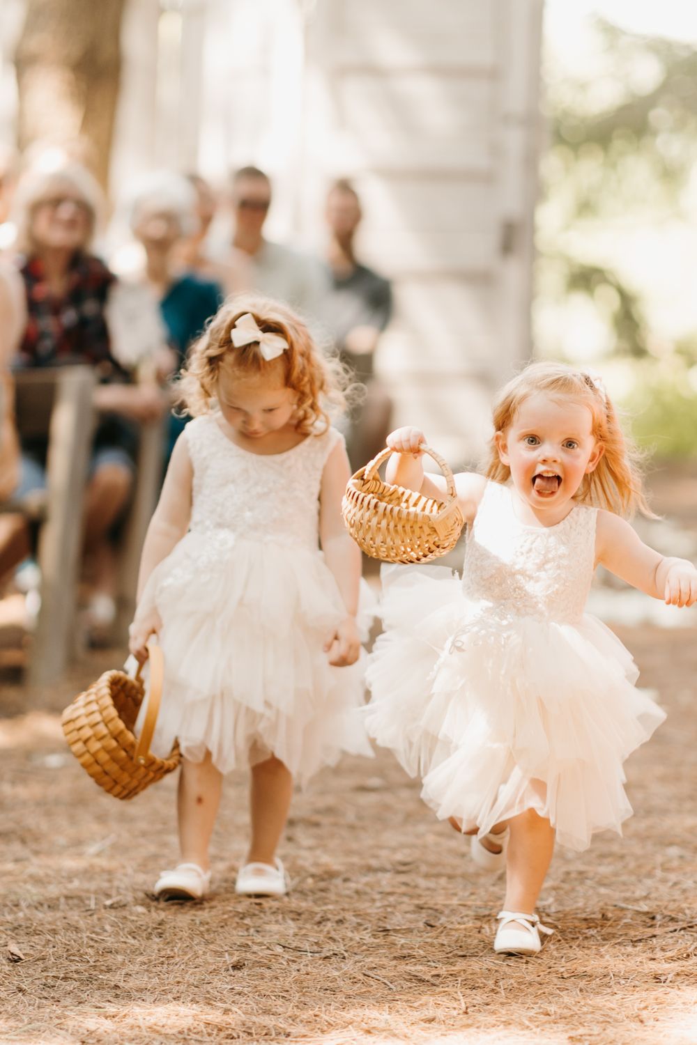 Two excited flower girls in white dresses carrying wicker baskets down outdoor wedding aisle.