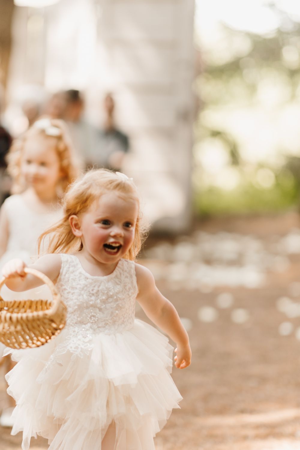 Joyful blonde flower girl in white tulle dress running down wedding aisle with wicker basket.