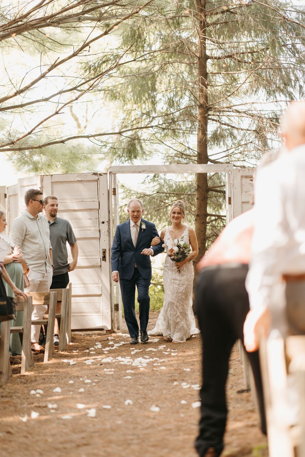 Bride walks down forest aisle with escort through vintage white doors at outdoor woodland wedding ceremony.