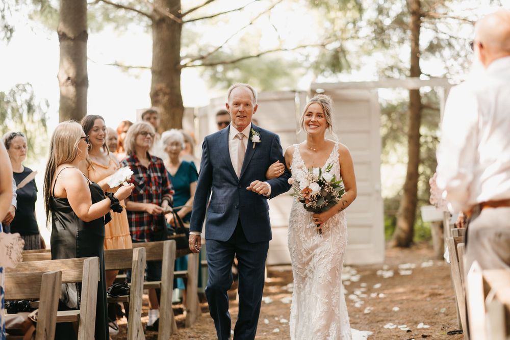 Smiling bride in lace gown walks aisle with escort past cheering guests at rustic outdoor wedding.
