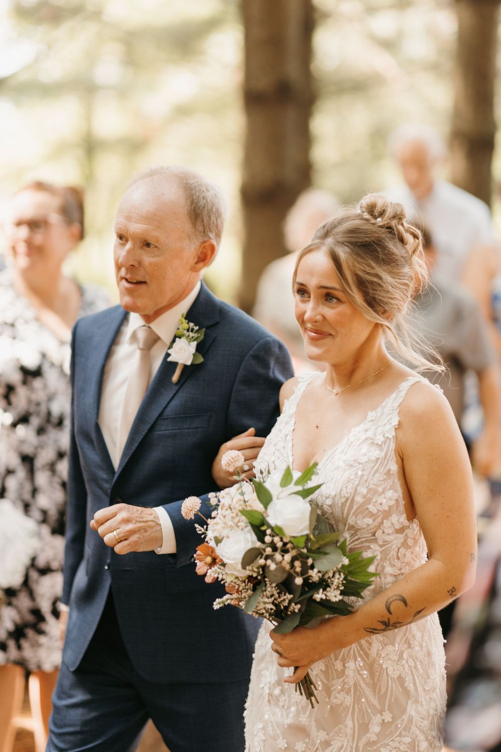 Emotional bride in lace gown holds bouquet while walking down aisle with older gentleman at wedding.