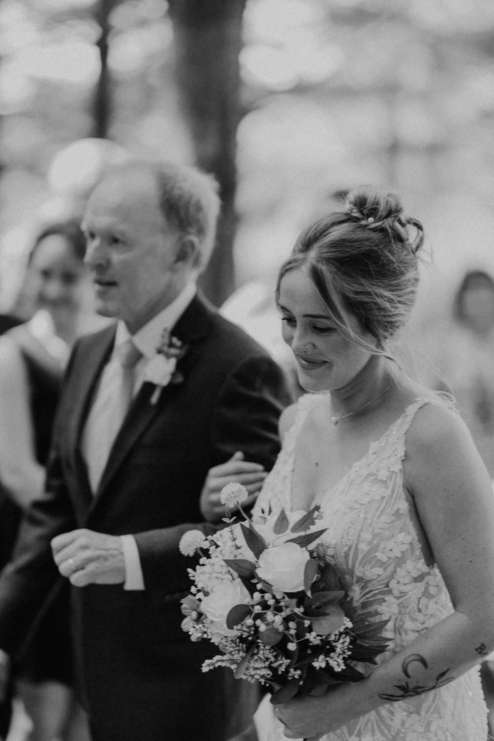 Black and white photo of bride holding bouquet walking down aisle with father at forest wedding.