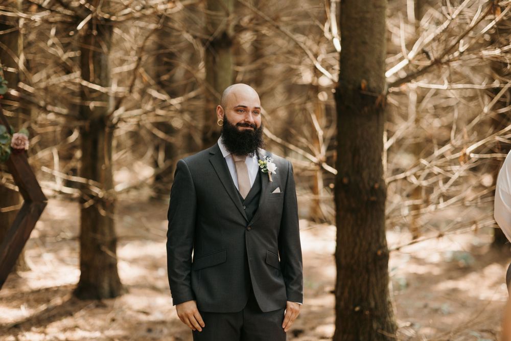 Bald bearded groom in dark suit stands among trees waiting at outdoor woodland wedding ceremony.