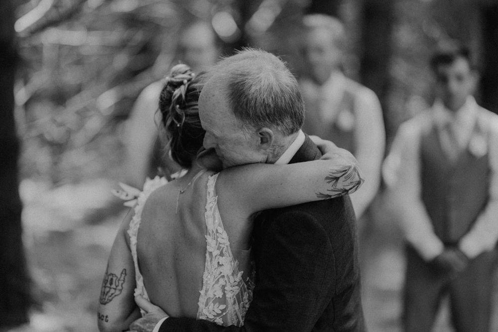 Touching black and white photo of bride and father sharing emotional hug at outdoor wedding ceremony.