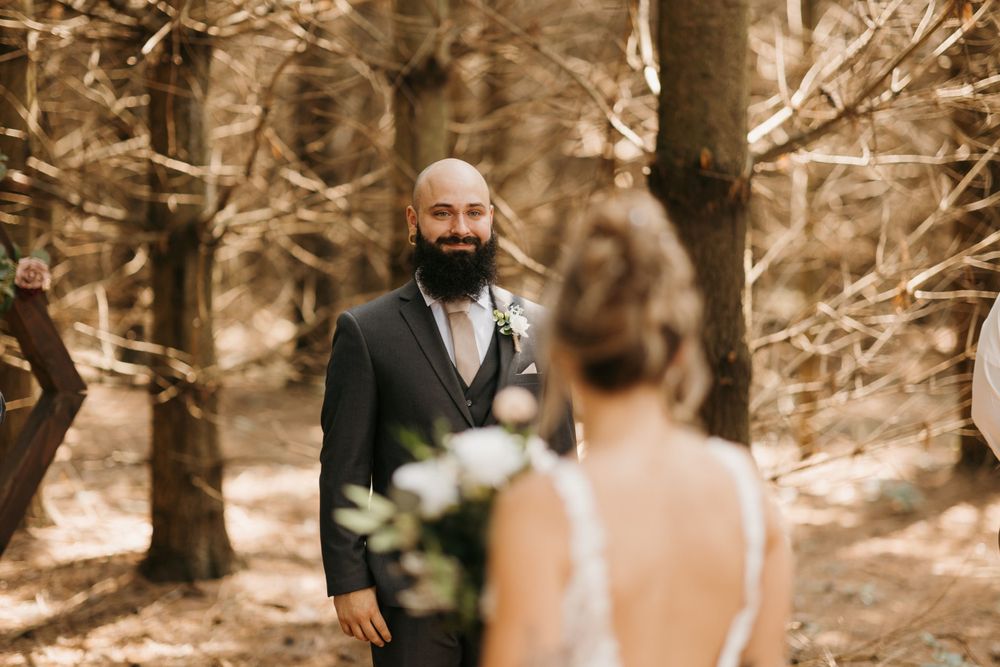 Bearded groom smiles seeing bride approach during outdoor woodland wedding ceremony among bare trees.