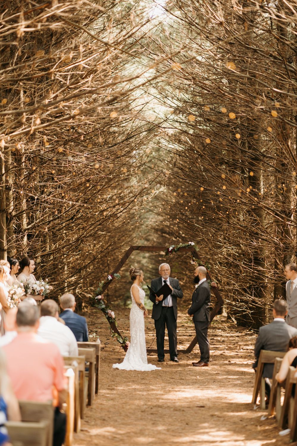 Couple stands at altar beneath golden arching tree canopy at enchanting outdoor forest wedding ceremony.