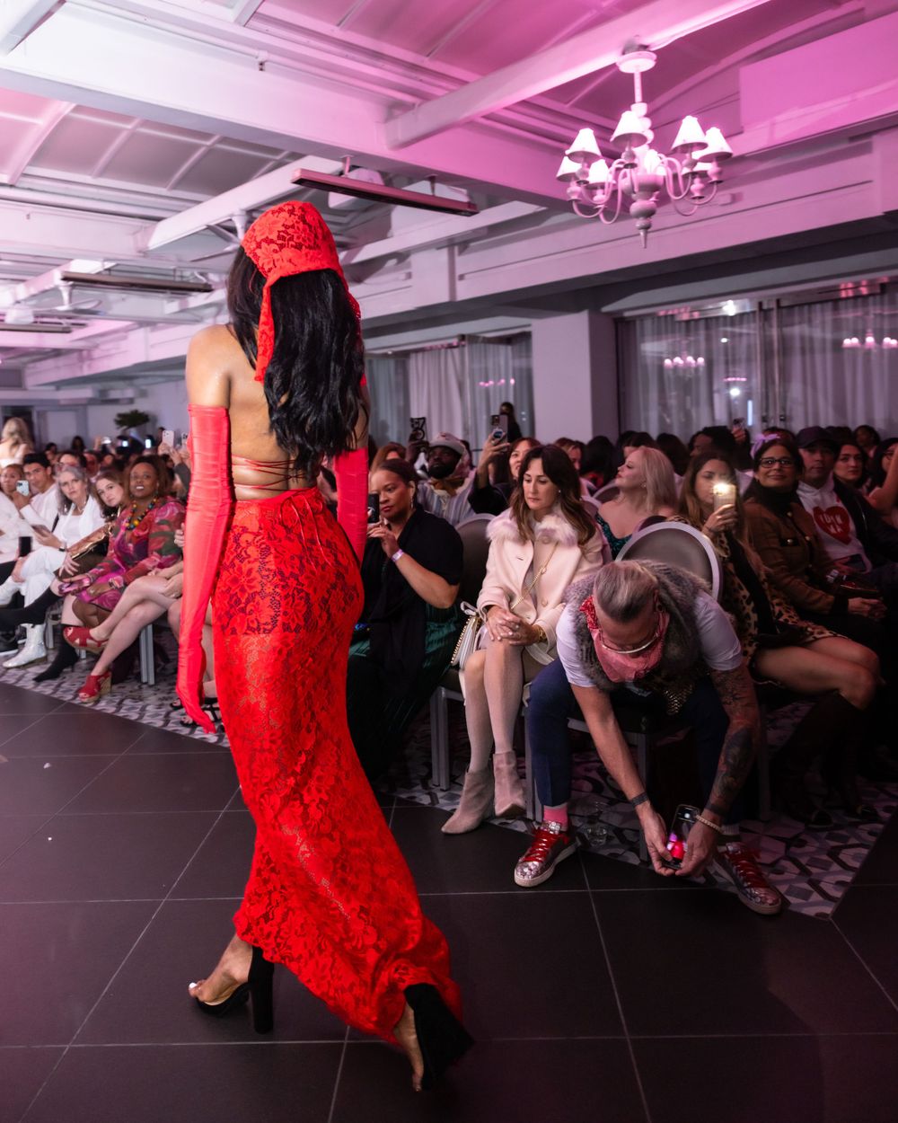 Model in red lace gown walks past audience during fashion show under pink lighting.