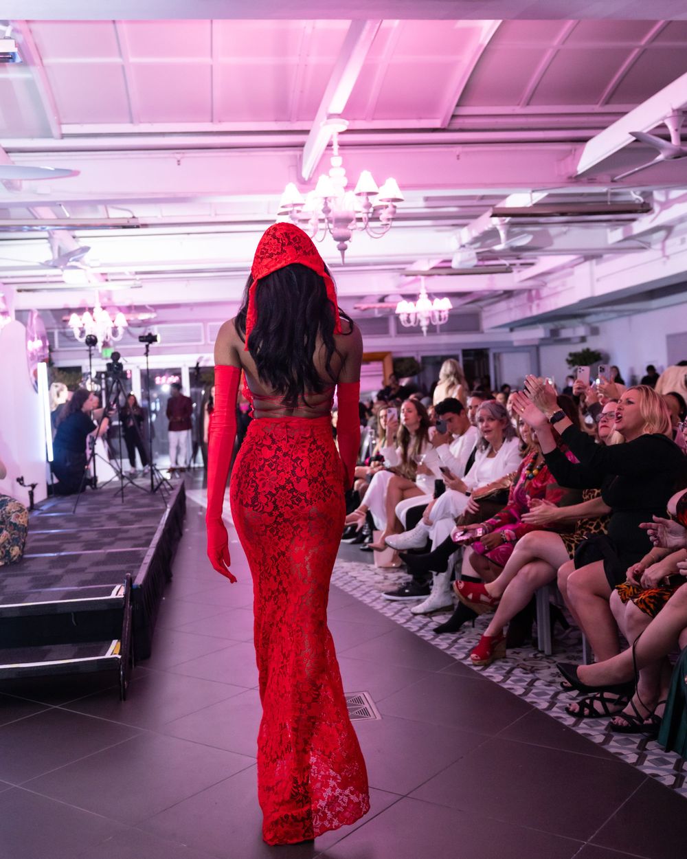 Back view of model in red lace gown walking runway at fashion show with pink ambient lighting.
