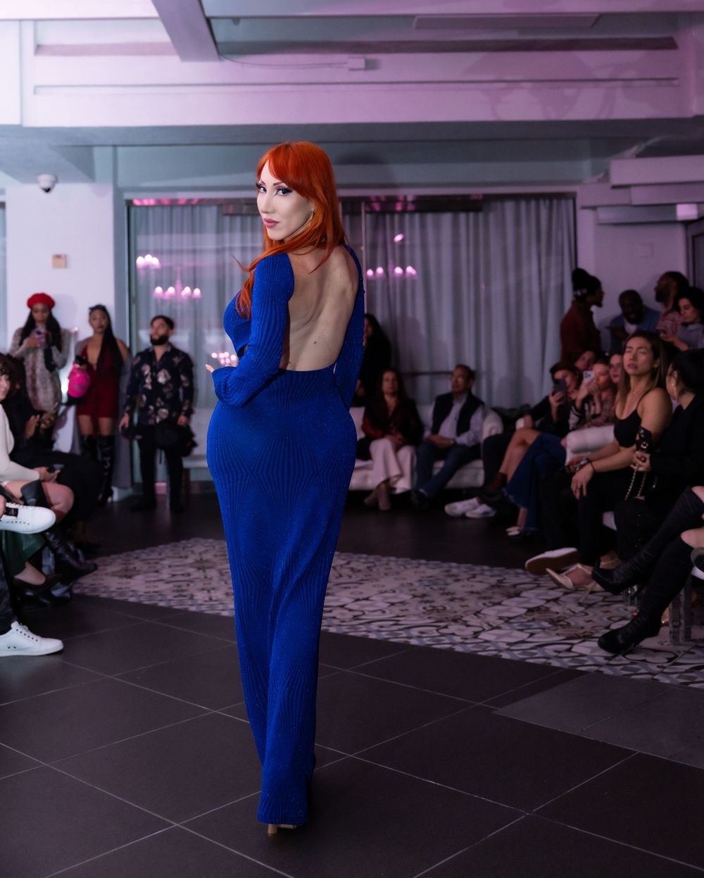 Fashion model wearing royal blue backless evening gown on runway with audience in background.